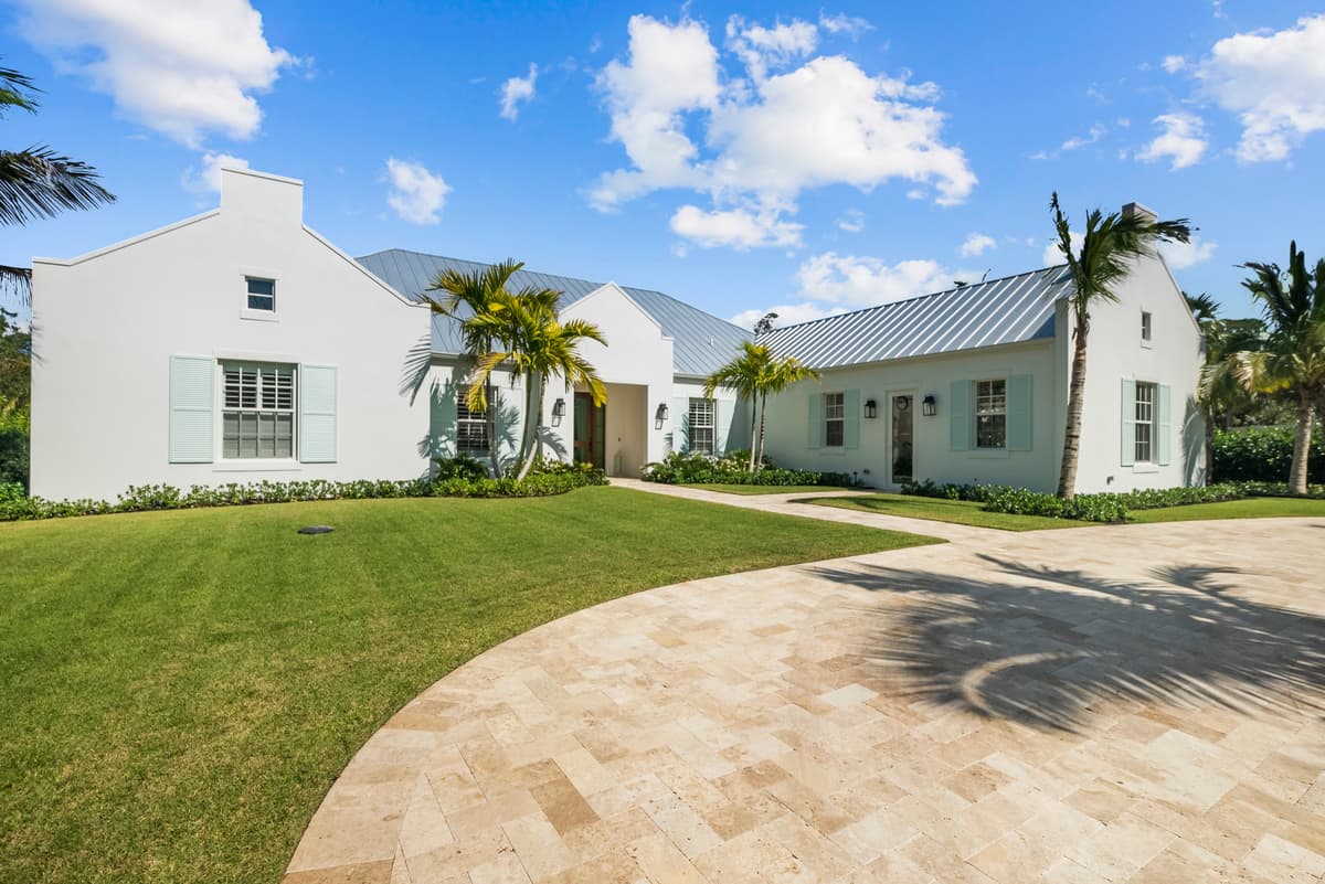 An estate residence in white stucco with Bermuda gables, pale shutters and a standing-seam roof, behind a curved travertine drive and lawn