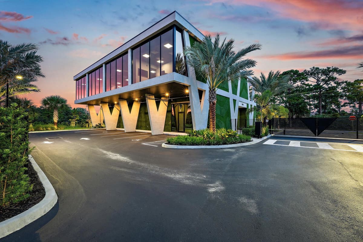 A two-story glass building raised on white V-shaped columns at dusk, palms either side of a private drive, in Hobe Sound